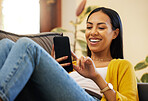 Hispanic woman using her smartphone while smiling and relaxing in a bright living room. A young female lying on a sofa scrolling on her cellphone at home using modern technology during lockdown