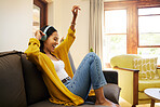 Woman listening to music on headphones while dancing in a bright living room. A happy smiling young female with eyes closed relaxing and sitting on a sofa at home using modern technology