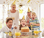 Cheerful generational happy family wearing party hats while celebrating little girl's birthday with confetti and cake at home. Excited kids, parents and grandparent enjoying special surprise event