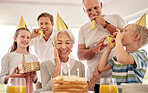 Senior woman celebrating her birthday with family at home, wearing party hats and blowing whistles. Grandma looking at birthday cake and looking joyful while surrounded by her grandkids, husband and son