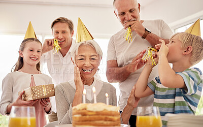 Senior woman celebrating her birthday with family at home, wearing party hats and blowing whistles. Grandma looking at birthday cake and looking joyful while surrounded by her grandkids, husband and son