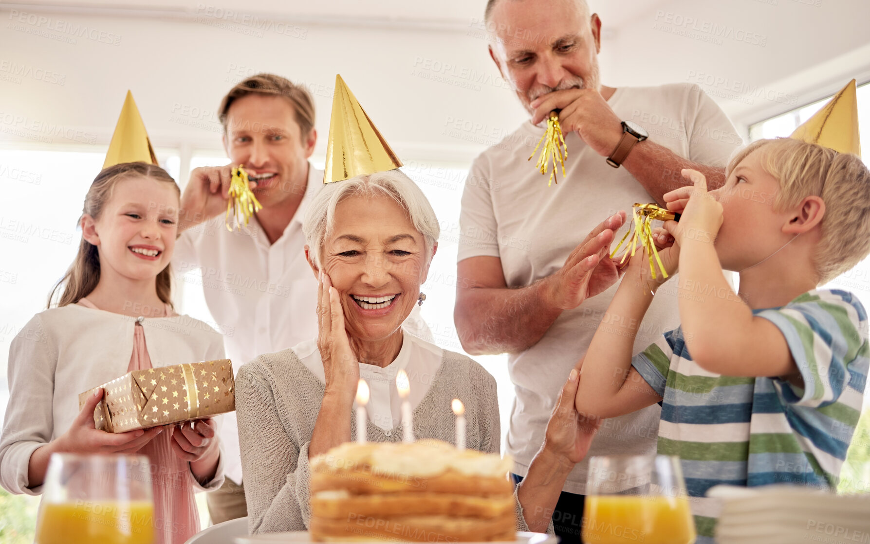 Buy stock photo Senior woman celebrating her birthday with family at home, wearing party hats and blowing whistles. Grandma looking at birthday cake and looking joyful while surrounded by her grandkids and and son