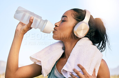 One fit young mixed race woman wearing wireless headphones taking a rest break to drink water from a bottle while exercising outdoors. Female athlete quenching thirst and cooling down while wearing a towel after running and training workout in nature