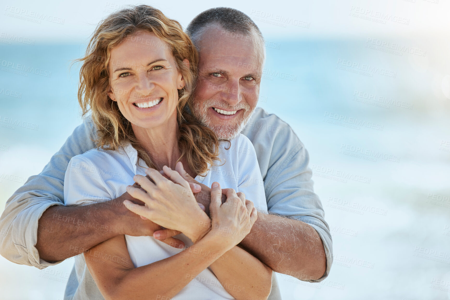 Buy stock photo Portrait of happy and loving retired mature caucasian couple enjoying a romantic date at the beach on a sunny day. Cheerful affectionate husband hugging his wife while enjoying seaside vacation