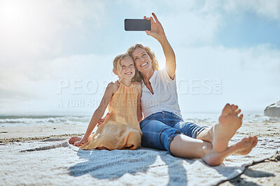 Buy stock photo Carefree mother and daughter taking a selfie while sitting on the beach. Happy little girl and grandmother smiling while taking a picture on a cellphone while on holiday. Mom and daughter bonding