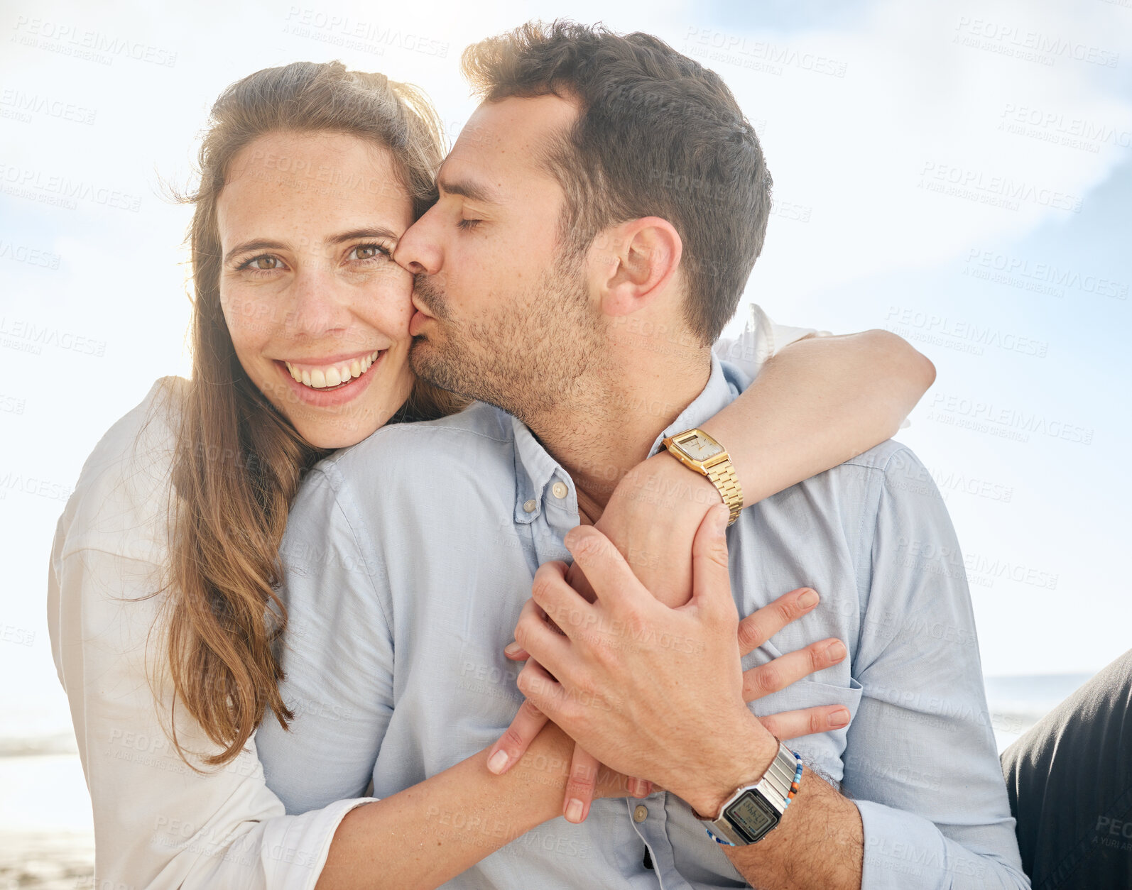 Buy stock photo Beautiful middle aged woman being kissed on the cheek by her husband while sitting outdoors on the beach. Affectionate married couple hugging on a romantic date, showing love to his wife outside