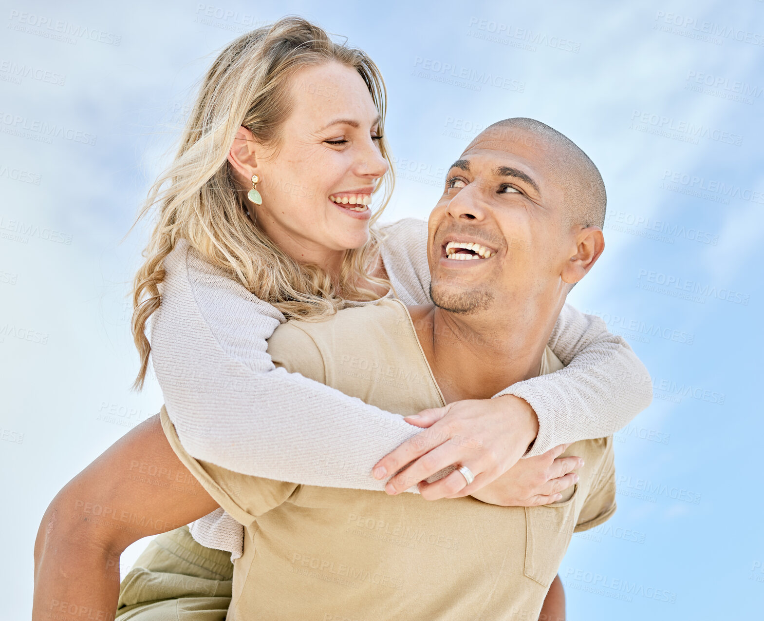 Buy stock photo Happy interracial couple enjoying a piggyback ride outside against a blue sky having fun and smiling at each other. Low angle of husband and wife together. Boyfriend carrying girlfriend on his back