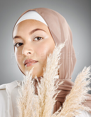 Studio portrait of one beautiful young muslim woman wearing brown headscarf posing with pampas wheat plant against grey background. Modest arab wearing makeup with face covered in traditional hijab