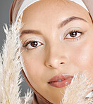 Closeup portrait of one beautiful muslim woman wearing brown headscarf posing with pampas wheat plant against grey studio background. Modest arab wearing makeup with face covered in traditional hijab