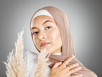 Studio portrait of one beautiful young muslim woman wearing brown headscarf posing with pampas wheat plant against grey background. Modest arab muslimah wearing makeup covered in traditional hijab