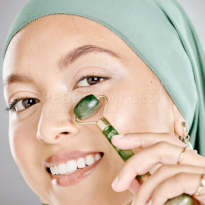 Buy stock photo Muslim woman smiling and using a facial roller on her skin. Beauty model doing her morning skin care routine with a jade roller. Young female using a gua sha product for healthy smooth skin.