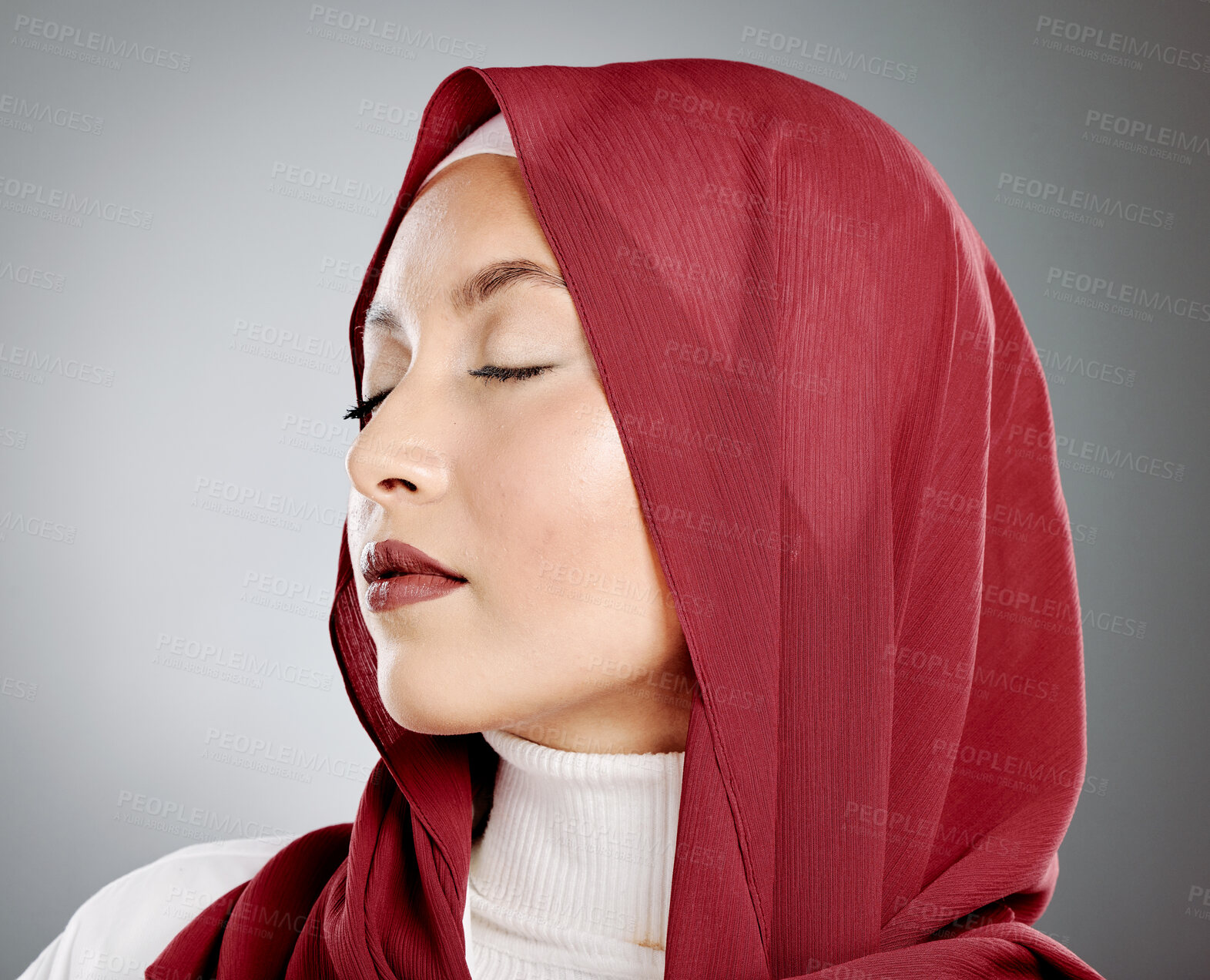 Buy stock photo Stylish young muslim woman in a red hijab with her eyes closed on a grey studio background. Middle eastern woman wearing makeup and a headscarf while feeling calm and showing the beauty in modesty