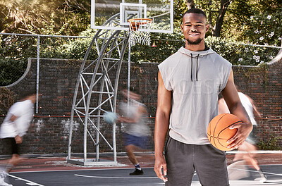 Portrait african american man standing with a basket ball on the court. Handsome male basketball player holding a sports ball while his friends and teammates play and practice in the background