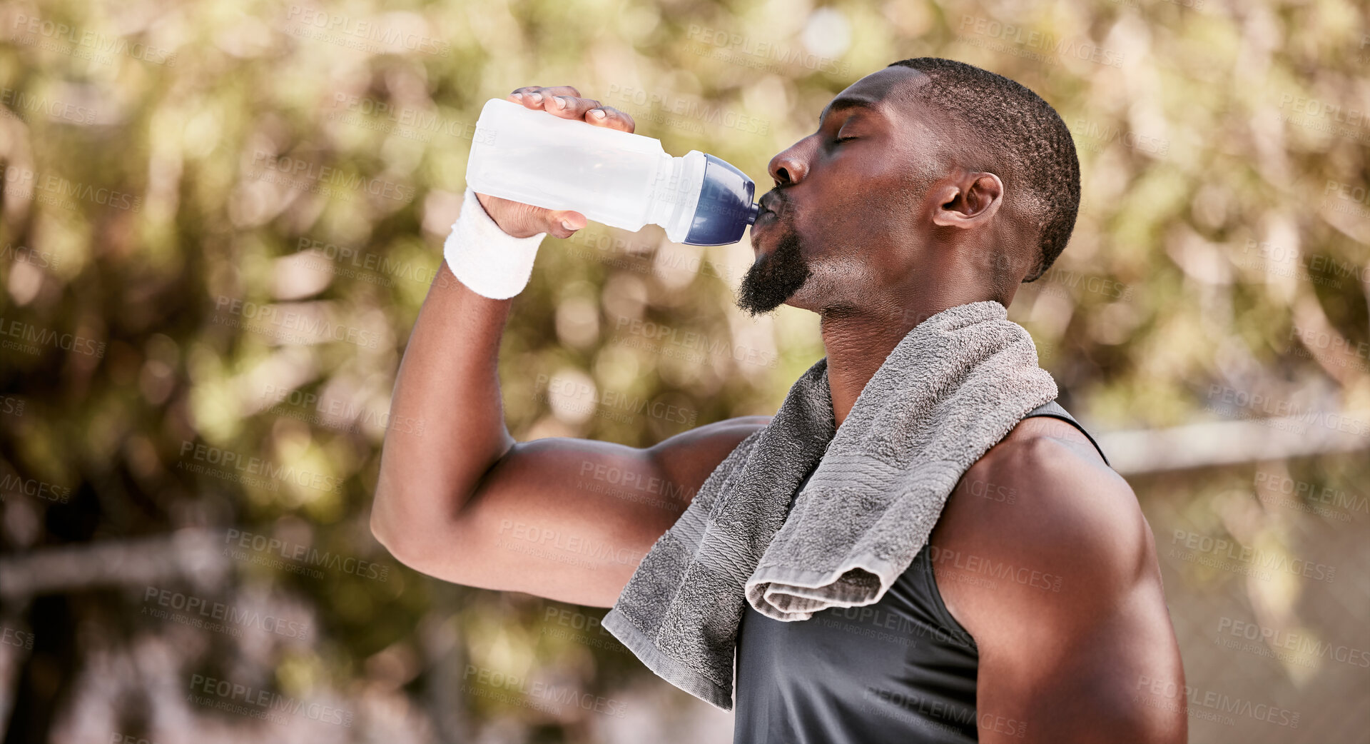 Buy stock photo Athlete drinking water from a bottle during an outdoor workout. Fit, athletic african american man taking break to hydrate and refresh while standing alone outside. Routine exercise for health
