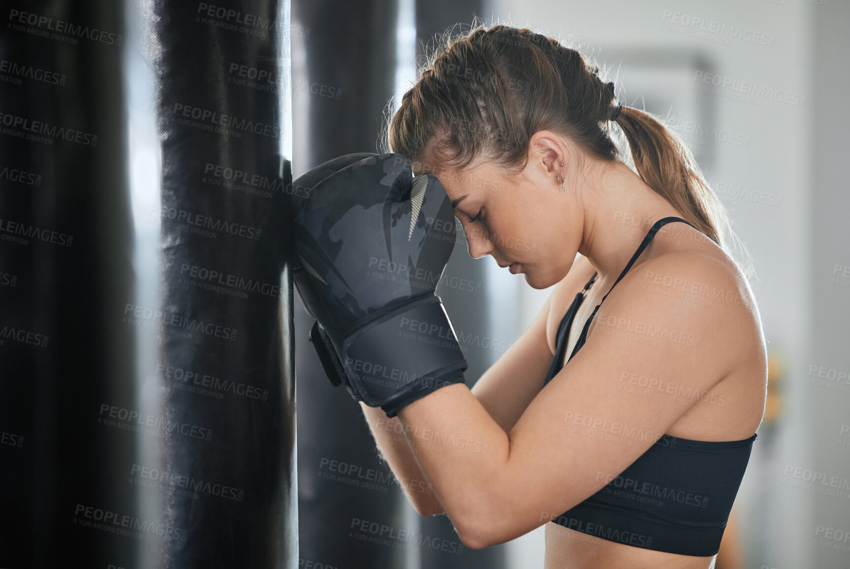 Buy stock photo Closeup of a tired and exhausted strong female athlete boxer, after a training session leaning on punching bag in the gym, side view. Fit and active woman taking a rest break after her exercise 
