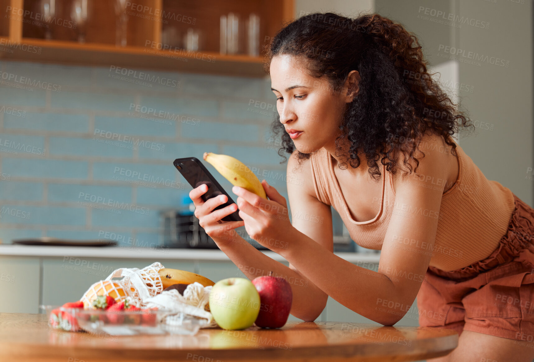 Buy stock photo Woman reading phone, researching a diet with fresh fruit while relaxing in a kitchen at home. Young female searching for a recipe, cleanse or detox online. Lady checking nutritional value of a banana