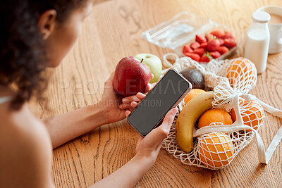 Buy stock photo Colorful, healthy and nutritional organic fruit groceries on table beside woman holding phone and looking online for smoothie recipes. Young lady eating fresh organic apple for wellness vegan diet
