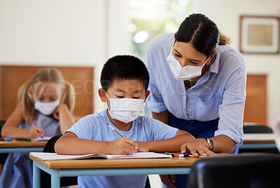 Buy stock photo Covid, education and learning with a teacher wearing a mask and helping a male student in class during school. Young boy studying in a classroom with help from an educator while sitting at his desk.