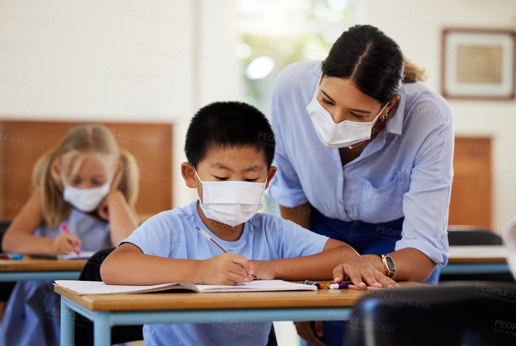 Buy stock photo Covid, education and learning with a teacher wearing a mask and helping a male student in class during school. Young boy studying in a classroom with help from an educator while sitting at his desk.