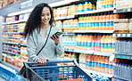 Grocery shopping, phone and woman using technology in a food store with groceries at a shop. Retail customer multitask in a supermarket walking with a mobile in a juice stock aisle and a happy smile