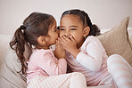 Two beautiful, little girls and pink pajamas. Whispering to each other, being playful on a pajama advertising set, sitting comfortably on a sofa, smiling in their diversity. Their hands on the mouth 