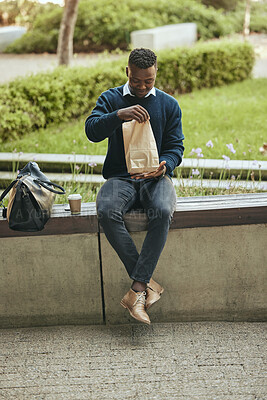 Buy stock photo Black business man on a lunch break in the city, sitting and eating a meal or sandwich in a brown paper bag. Hungry worker relaxing with a tasty snack, food on the go for a professional entrepreneur