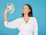 Money, wealth and rich woman holding fan of cash and cooling herself, ready to blow or spend it all. Female lottery winner embracing luck, success and win in the studio on a blue background.