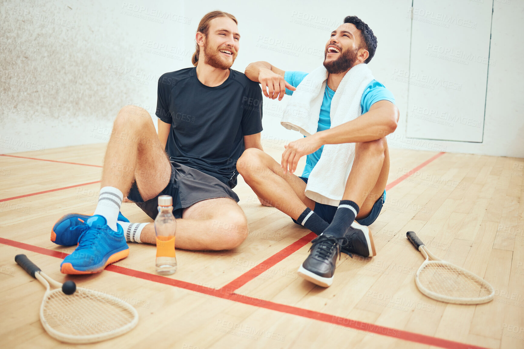 Buy stock photo Two young athletic squash players taking break after playing court game. Full length of laughing mixed race and caucasian athlete sitting together after training practice in sports centre. Sporty men