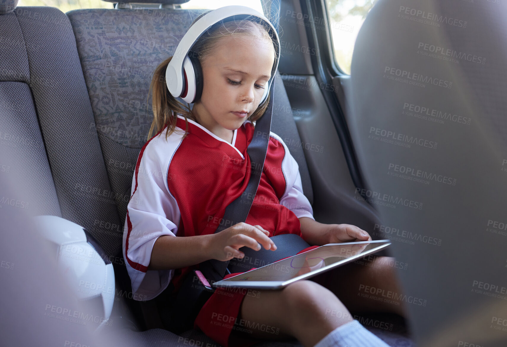 Buy stock photo Child with tablet, headphones and safety belt in the car going home after soccer match, game or practice. Young girl in sports gear playing educational games online on tech device after training