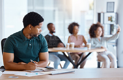 Buy stock photo Lonely, alone and African businessman in the office working on a project while colleagues take selfie. Reject, sad and antisocial professional employee sitting at his desk as outcast in the workplace