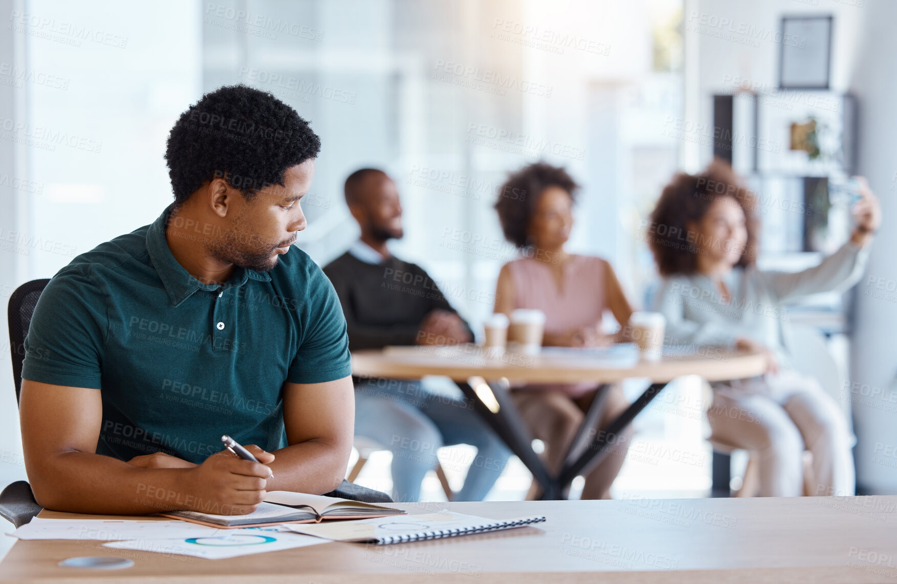 Buy stock photo Lonely, alone and African businessman in the office working on a project while colleagues take selfie. Reject, sad and antisocial professional employee sitting at his desk as outcast in the workplace