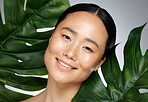 Face, beauty and asian woman with monstera leaf on gray studio background. A beautiful Japanese woman with healthy, glowing and flawless skin, skincare routine and makeup with organic plant cosmetics