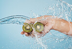 Hands, beauty and kiwi in a studio with splash for skincare, shower and organic treatment against a blue background. Hand, water and fruit with woman wellness model grooming, hygiene and cleaning