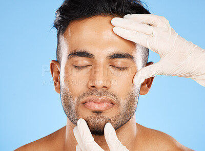 Buy stock photo Cosmetic, plastic surgery and face of young man, for skincare and white gloves on blue studio background for dermatology care. Makeup, Indian male and skin rejuvenation for wellness and collagen.