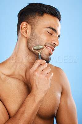 Buy stock photo Skincare, beauty and man with a jade roller for face massage against a blue studio background. Wellness, health and happy cosmetics model with a tool for facial treatment and smile for relax