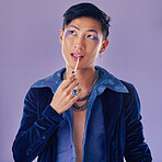 Beauty, makeup and lgbt Asian man in studio on purple background using cosmetics, beauty products and lipstick. Creative, fashion and queer or lgbtq male model with lip gloss, eyeshadow and style