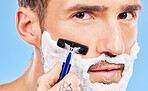 Grooming, cream and man shaving his face with a razor for wellness, skincare and health against a blue studio background. Beauty, young and portrait of a model with foam on beard for hygiene