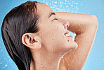 Shower, water and face of a woman cleaning body to relax for wellness against a blue studio background. Hair care, happy and Asian model washing for grooming, hygiene and health with a smile