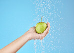 Green apple, hands and water splash for healthy lifestyle, vegan diet and nutrition on studio blue background. Palm holding fruits under clean running water for wellness, detox snack and vitamins 