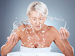 senior woman, water splash and skincare with cleaning, wellness and face wash against a grey studio background. Elderly model, beauty and facials washing with liquid, hands and beauty treatment