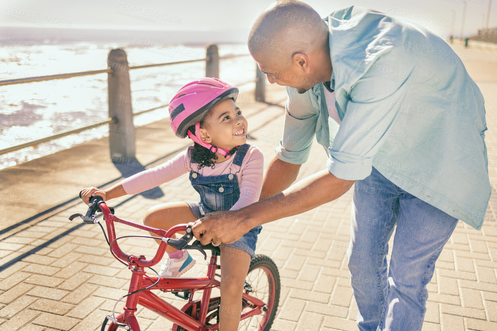 Buy stock photo Father teaching his child to ride a bike at the beach while on a summer vacation, holiday or adventure. Happy, learning and man helping his girl kid with her bicycle while bonding in nature by ocean.