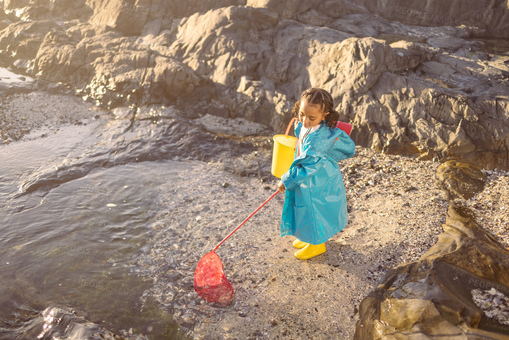 Buy stock photo Girl, beach and fishing with net in rock pool, water or ocean with boots, bucket and coat in sunshine. Black child, sea and outdoor for fish, marine life or animal in nature, learning and adventure