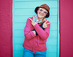 Portrait, peace and funky with a senior woman outdoor standing against a blue door and red wall background. Glasses, hands and hip hop with a happy mature female doing a hand sign or gesture outside