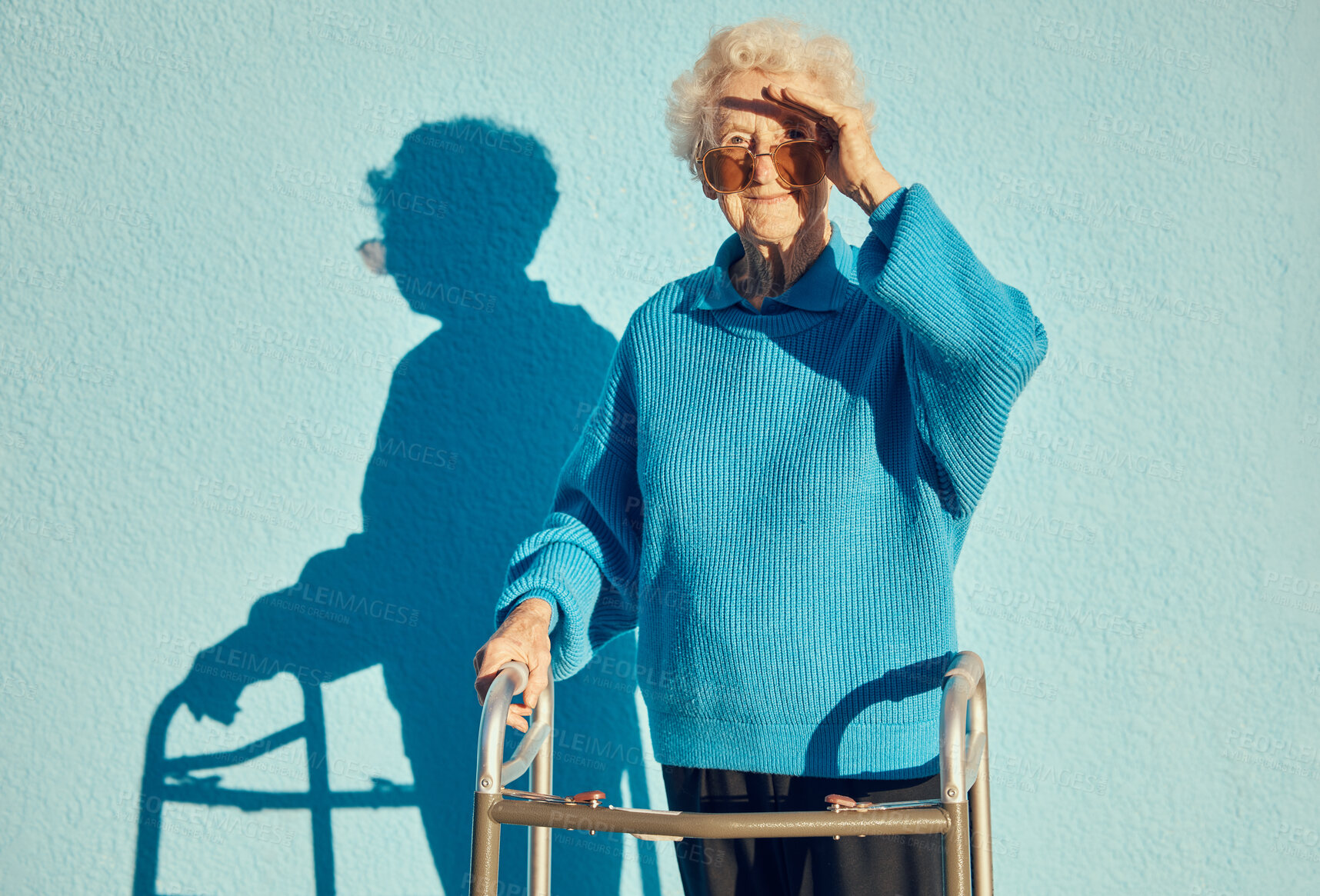 Buy stock photo Portrait, shadow and disability with a senior woman on a blue wall background while holding a mobility walker outdoor. Health, handicap and fashion with a mature female standing outside alone