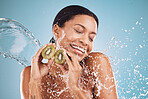 Woman, kiwi and water splash for skincare, cosmetics and natural beauty on blue studio background. Wet, young female and black girl fruit for wellness, clear or smooth skin for health, smile or relax