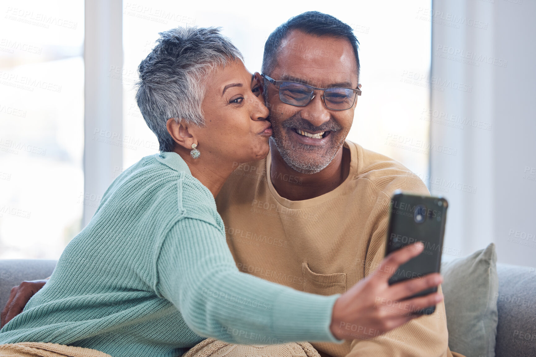 Buy stock photo Phone, selfie and kiss with a senior couple posing for a picture in the living room of their home together. Smartphone, kissing and social media with a mature man and woman taking a photograph