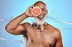 Skincare, portrait and senior man with grapefruit in studio for beauty, water splash and wellness on blue background. Face, beauty splash on fruit product by elderly man for facial, skin and cleaning