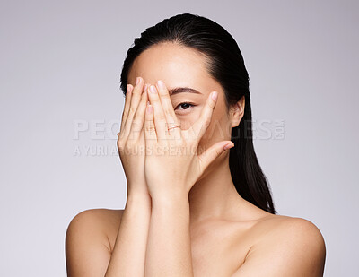 Buy stock photo Shy, beauty and woman hiding face in studio on a gray background. Makeup, skincare cosmetics and portrait of introvert, young and Asian female model peeking through her hands after facial treatment.