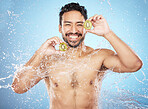 Face portrait, water splash and man with kiwi in studio isolated on a blue background. Cleaning, skincare and hygiene of happy male model washing fresh fruit for vitamin c, nutrition and healthy diet