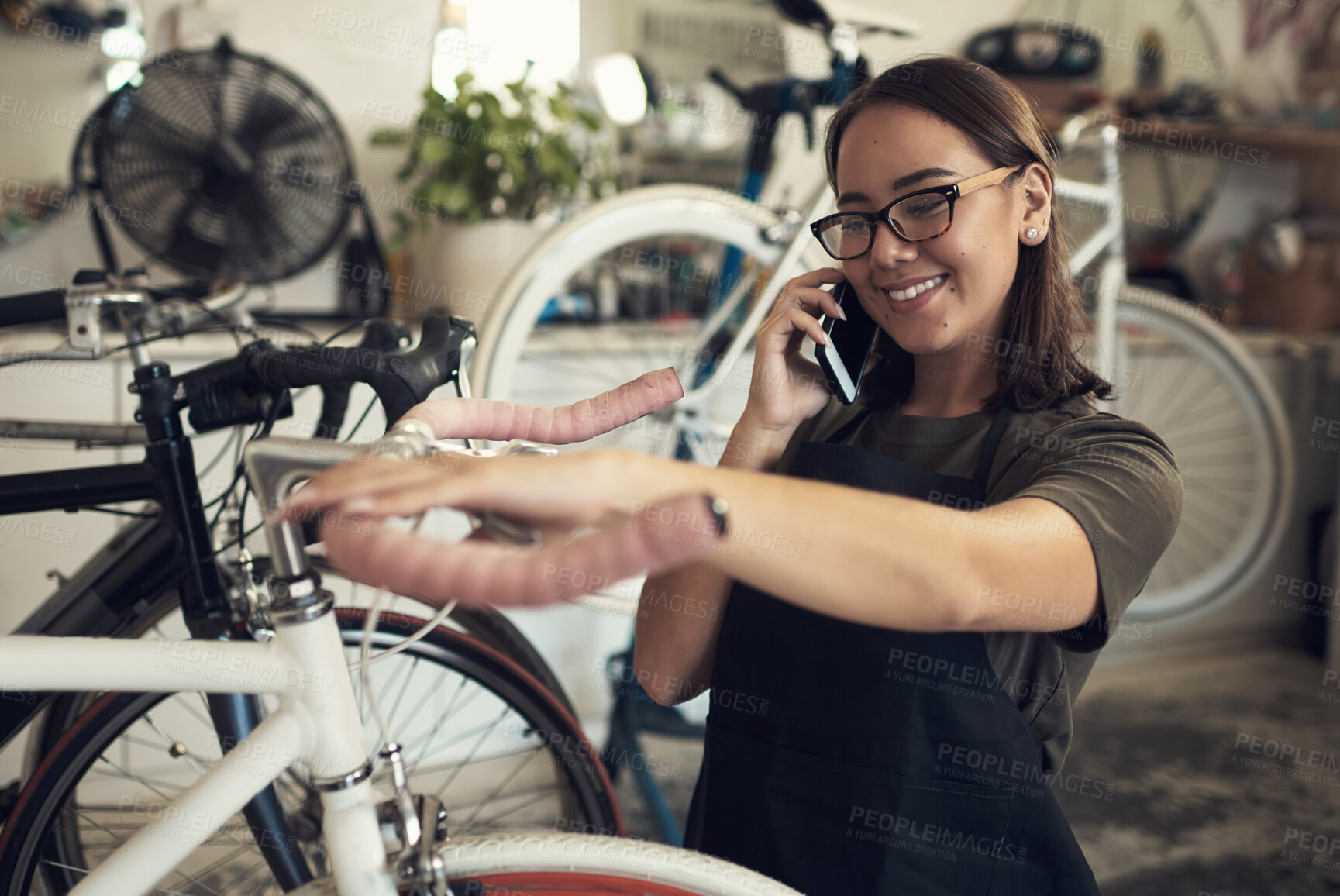 Buy stock photo Shot of an attractive young woman standing alone in her shop and repairing a bicycle wheel while using her cellphone